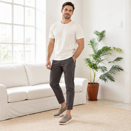 Man in a white T-shirt and charcoal pants wearing taupe XE5813 knit slip-on shoes in a living room.